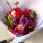 The Dahlia Delight Bouquet - Pop of Colour by Flowers on Norton St features vibrant seasonal dahlias in red, pink, purple, and cream. Wrapped in white paper, these lush blooms are perfect for weddings and showcase various sizes and petal arrangements elegantly held by hand.