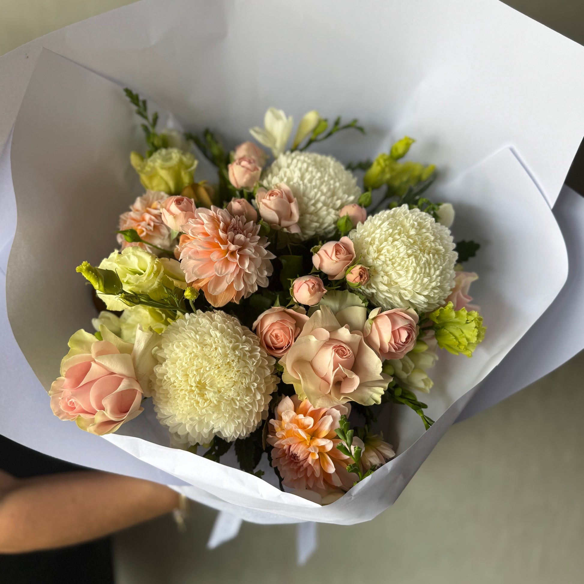 The Little Sweetheart Posy by Flowers on Norton St presents in-season flowers, highlighting white and peach roses, chrysanthemums, and dahlias tastefully wrapped in crisp white paper.