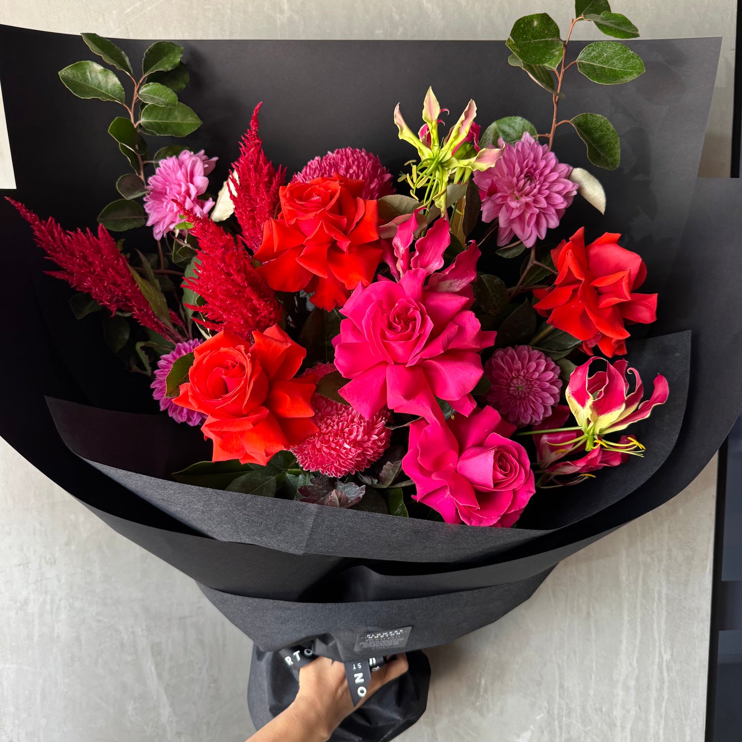 The Florist Choice - Red Nina Bouquet by FlowersonNortonSt boasts vibrant Red Nina and Colombian roses with purple dahlias and green leaves, beautifully wrapped in black paper.