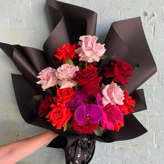 The Rose Phal Ruffle Bouquet from Flowers on Norton St features a delightful arrangement of mixed-colored roses in red and pink hues, paired with vivid Phalaenopsis orchids, all beautifully wrapped in a ruffle wrap against the backdrop of a white brick wall.