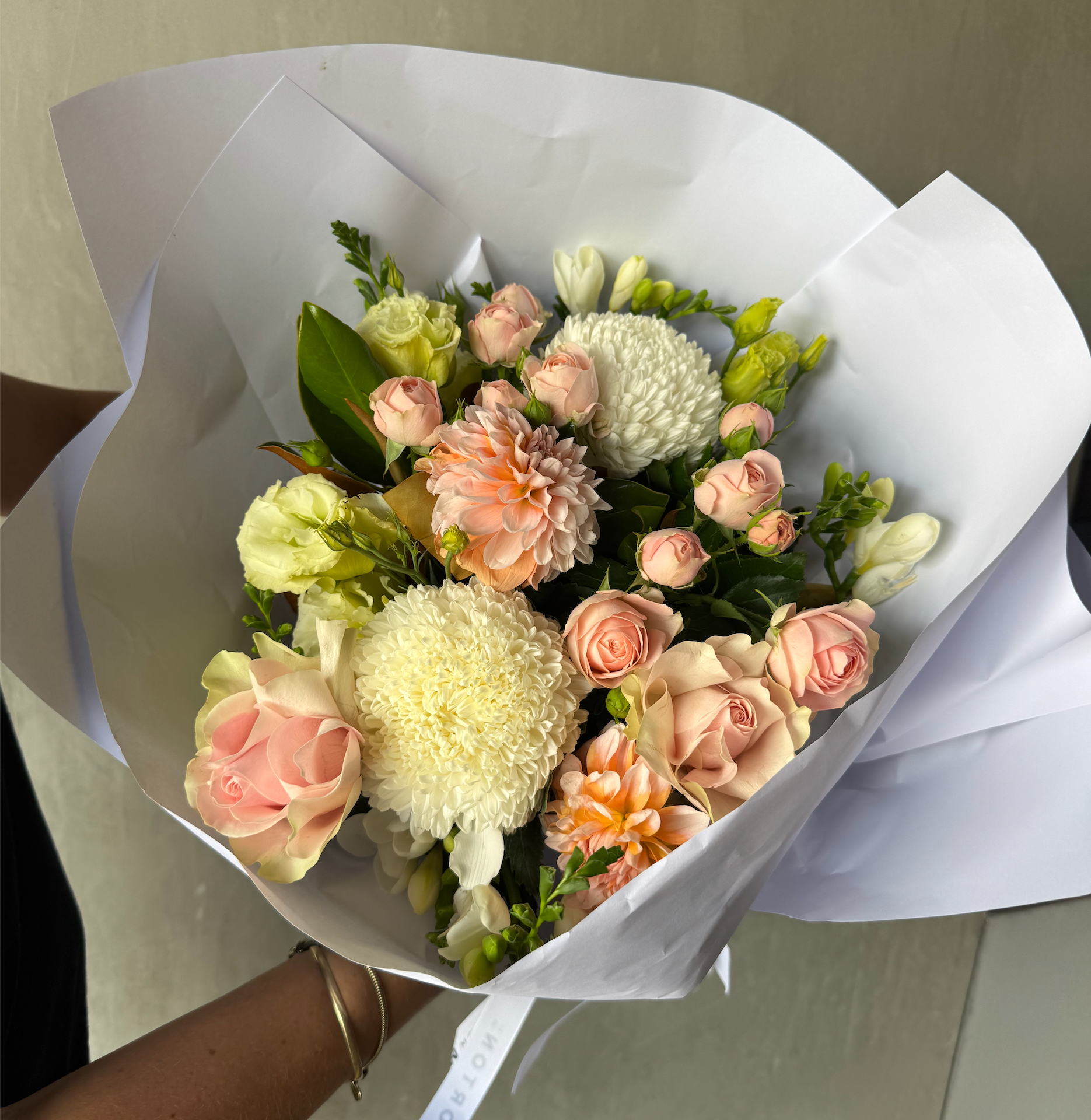 The Little Sweetheart Posy by Flowers on Norton St features white chrysanthemums, peach roses, and pink spray roses with greenery. Wrapped in white paper, it's expertly crafted with florist-selected in-season flowers for a natural, elegant look against a muted background.