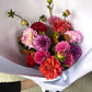 The Dahlia Delight Bouquet - Pop of Colour from Flowers on Norton St, featuring vibrant red, pink, purple, and orange dahlias wrapped in crisp white paper, is ideal for weddings. A hand gently holds this lively arrangement of fresh seasonal blooms.