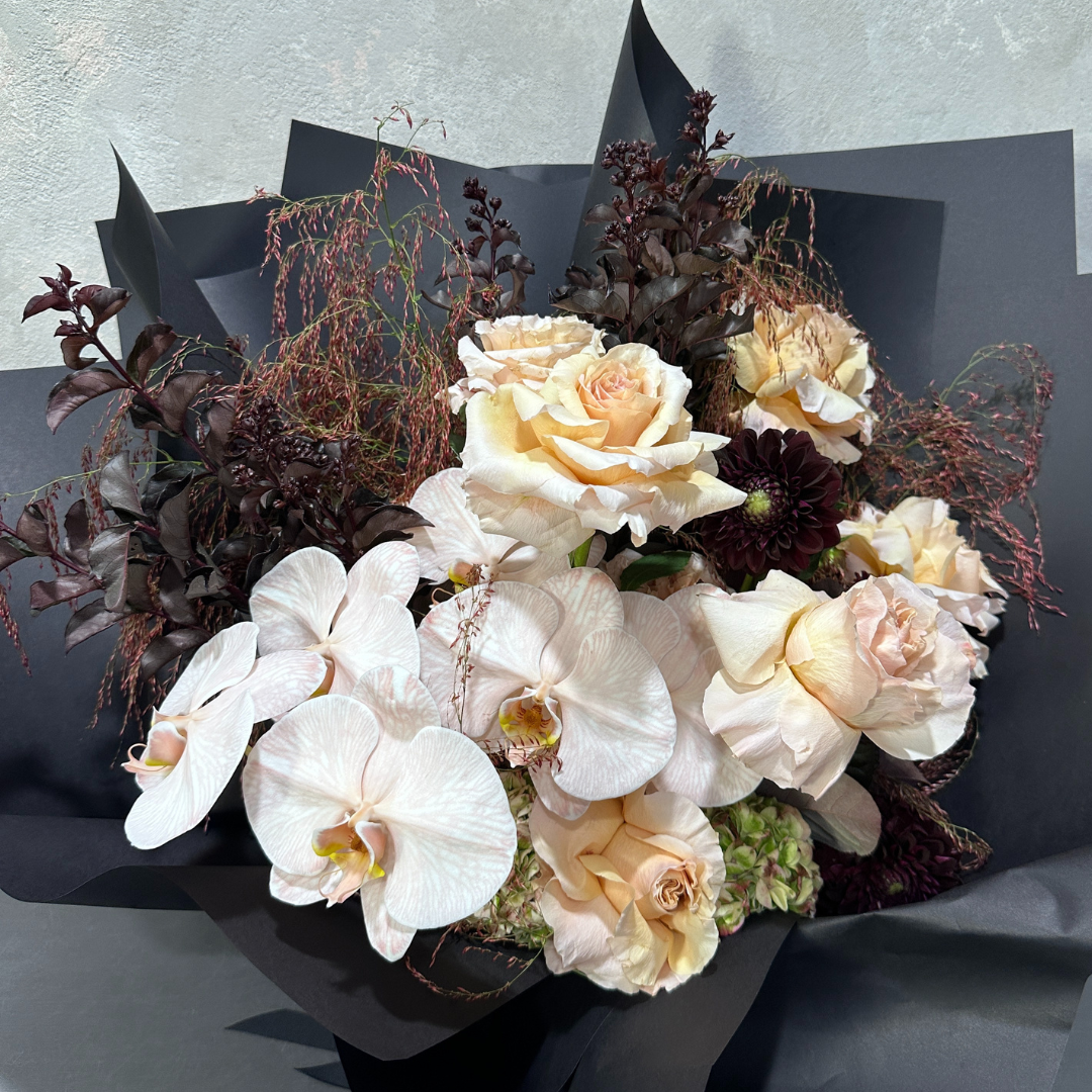 Introducing the Champagne Delight by Flowers on Norton St: an exquisite floral arrangement showcasing Colombian rose shimmer, phalaenopsis orchids, and dark purple foliage, all elegantly wrapped in black paper. The contrast between light and dark elements creates a stunning visual effect.