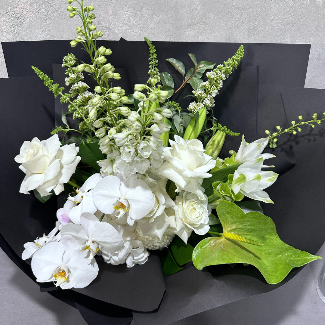 The "Under the Limelight" bouquet from Flowers on Norton St features a stunning combination of white roses, lilies, and orchids, expertly arranged with greenery and lime anthuriums against a sleek black wrapping. The bouquet is elegantly displayed on a light gray textured surface.