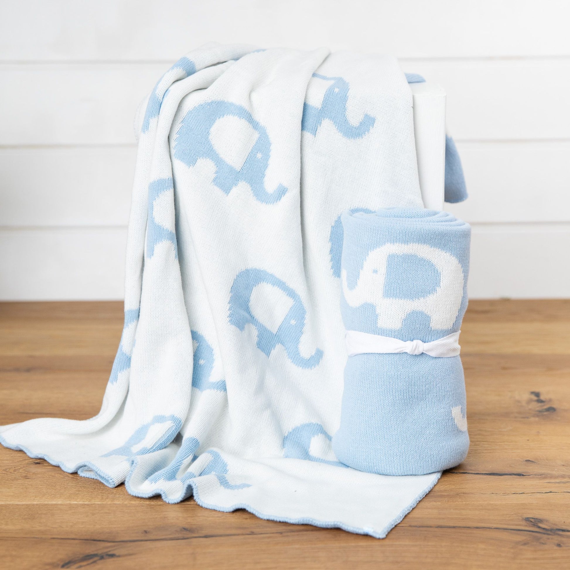 A cuddly Ellie Baby Blanket by FlowersonNortonSt, featuring a white base with blue elephants, drapes over a chair. It is accompanied by another Ellie Baby Blanket rolled up and tied with a white ribbon, both set gracefully on a wooden floor against a pristine white backdrop.