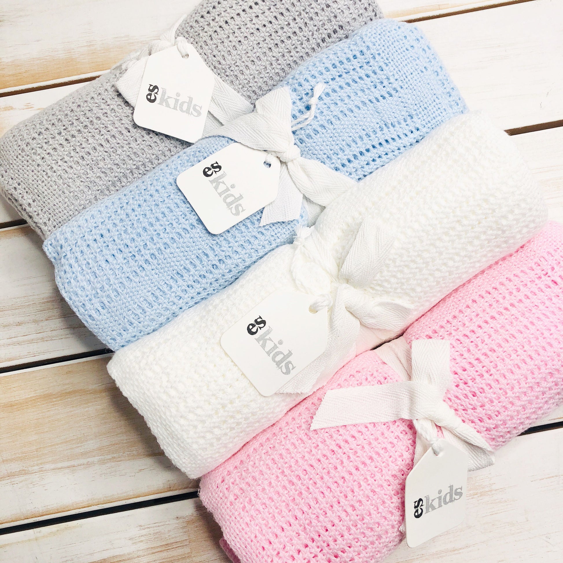 On a wooden surface, four rolled Baby Blankets by FlowersonNortonSt are displayed in gray, blue, white, and pink. Each blanket features ribbon ties and tags reading "Baby Kids." Crafted for snuggling, these soft blankets offer a cozy experience for your little ones.