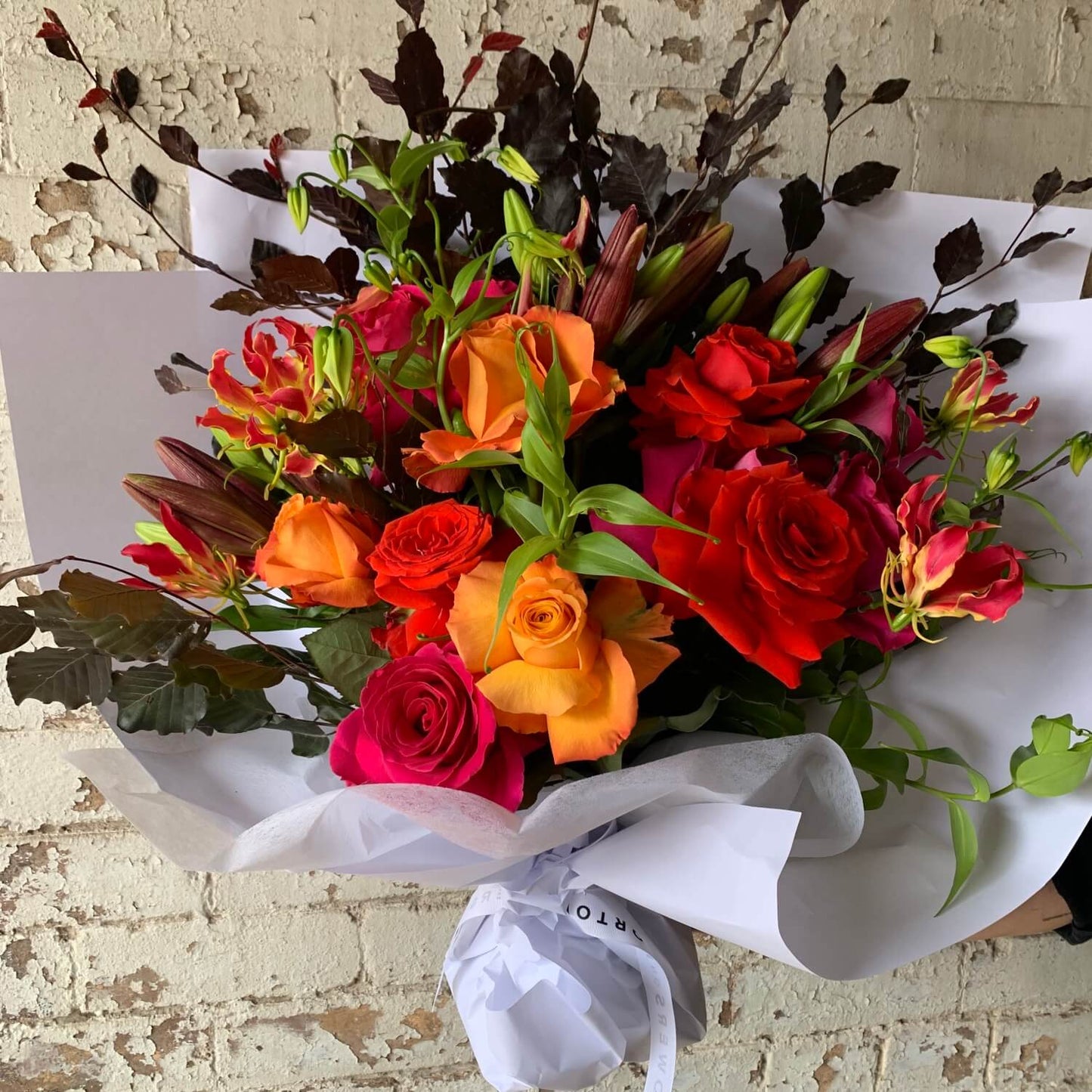 The "Floral Decadence" bouquet from Flowers on Norton St presents a lively winter assortment of red, orange, and pink roses and lilies, accentuated by rich dark foliage. Wrapped in crisp white paper, this striking arrangement is elegantly displayed against a textured, light-colored brick wall.