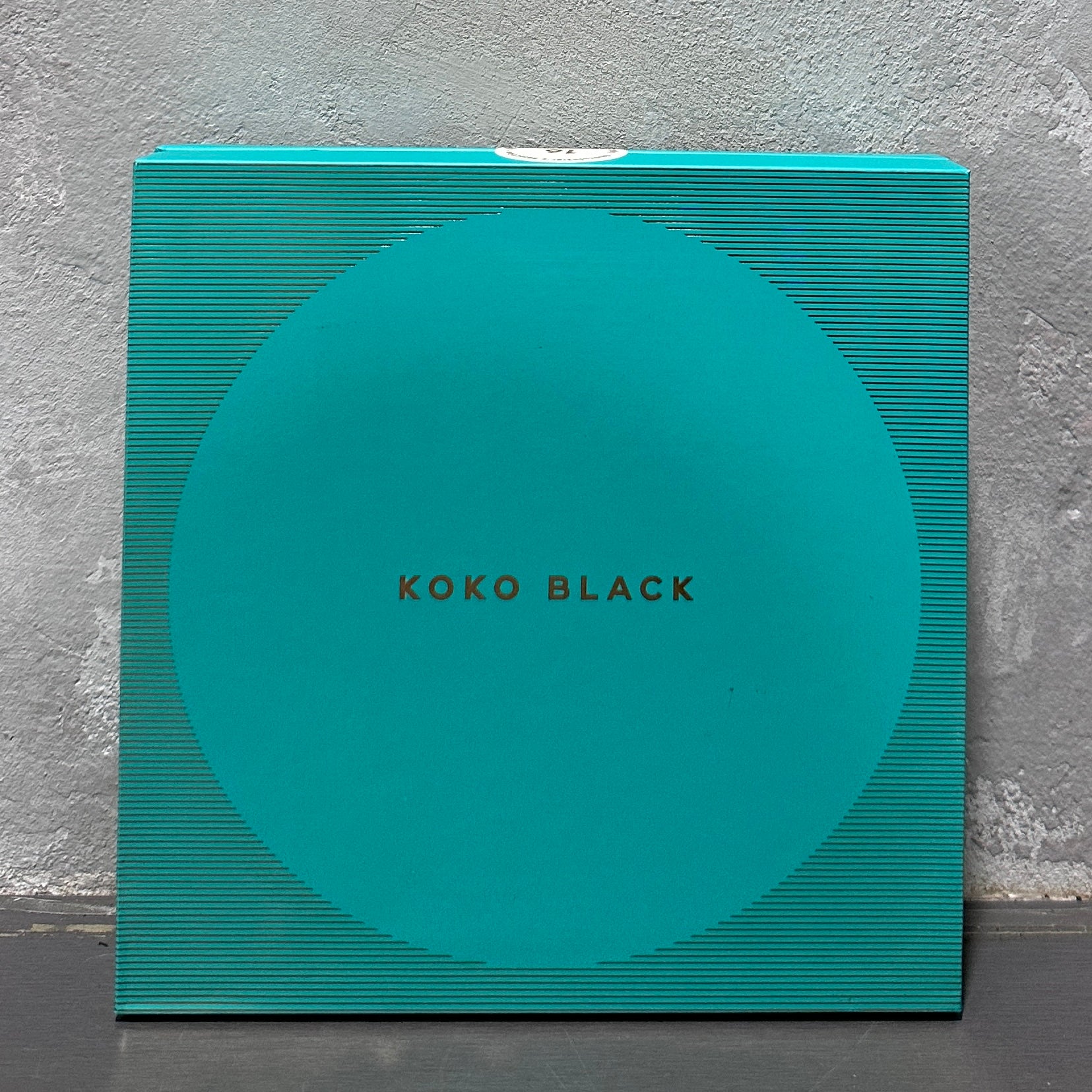 A large chocolate box from Flowers on Norton St, featuring a teal color with subtle textured stripes and gold lettering on the front, is set against a gray textured wall.