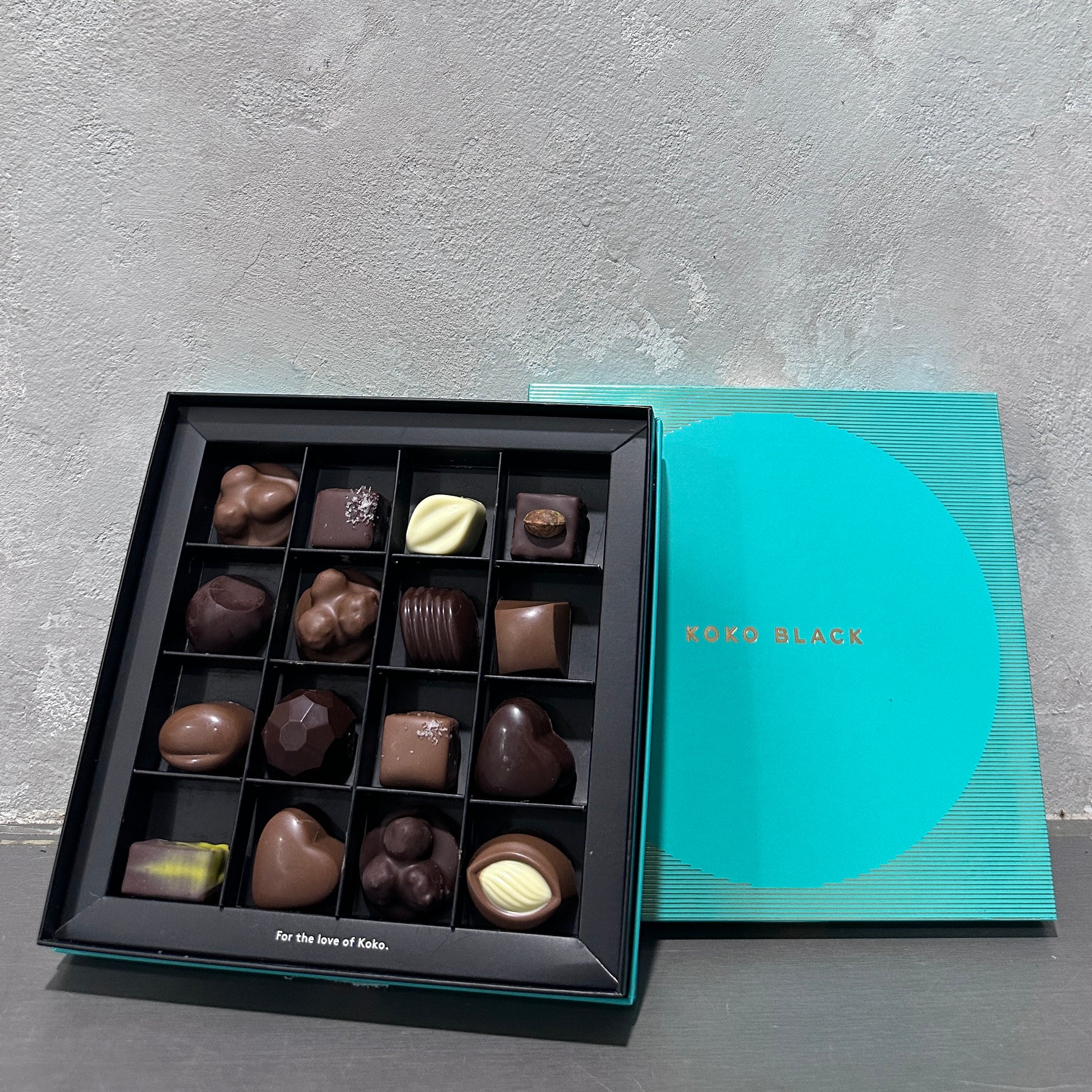 An elegant teal "Koko Black Chocolate Box Large" from Flowers on Norton St is displayed, partially opened, showcasing an assortment of chocolates with various shapes and designs on the gray textured surface.