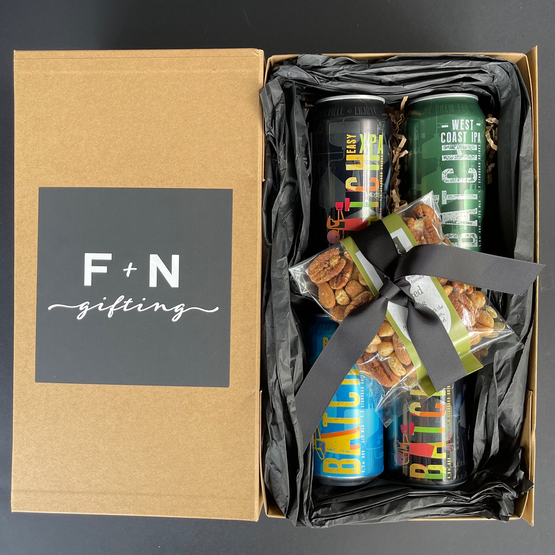 Gift box featuring the "Cans of Craft Beer Gift Box" from Flowers on Norton St, which includes three colorful cans of craft beer and a packet of spiced nuts, all tied with a sleek black ribbon. The slightly open lid showcases "F+N gifting" in crisp white against a dark, plain background. Ideal for adding a touch of elegance to any occasion.