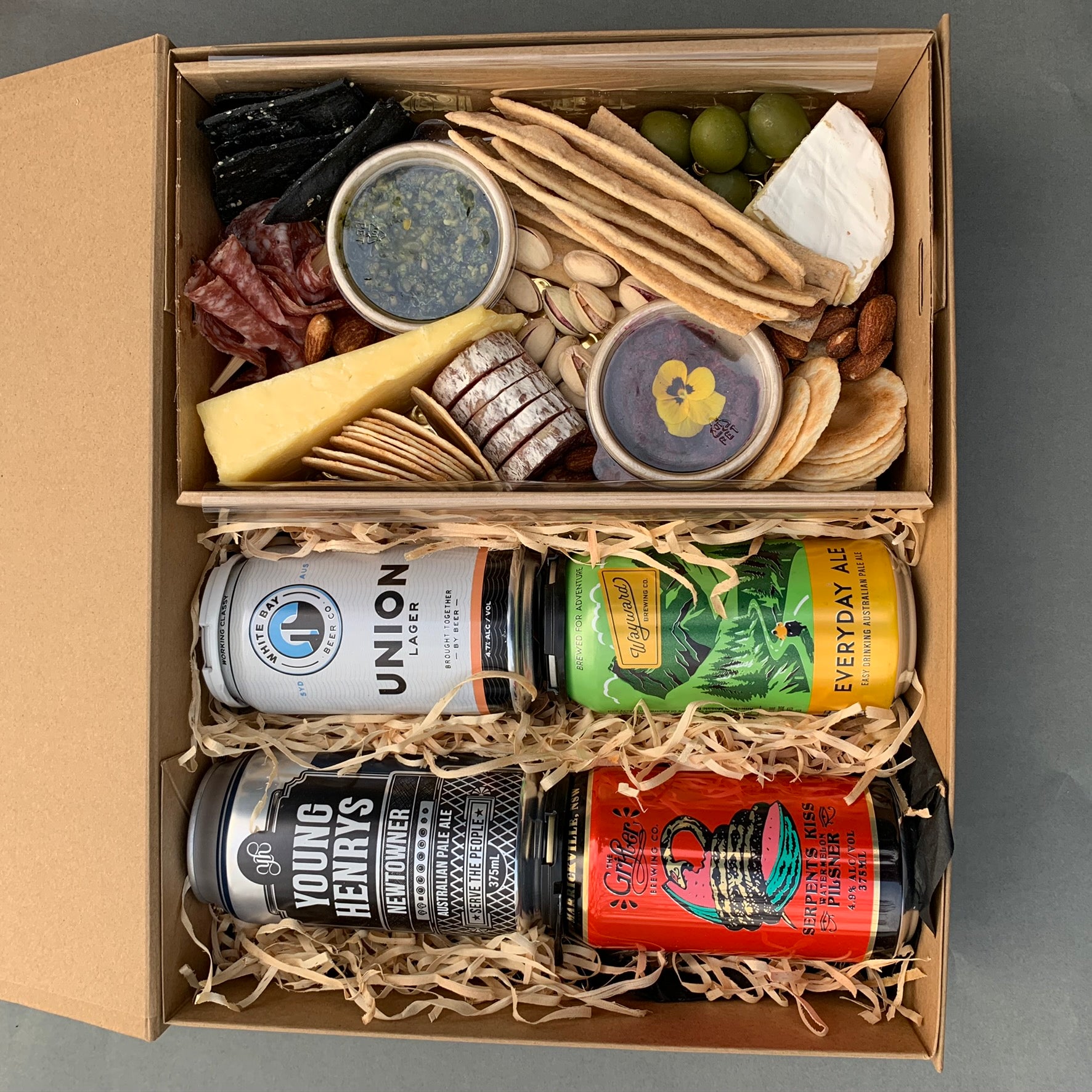 A gourmet gift box from Flowers on Norton St, known as the "Cans of Craft Beer Gift Box," includes three cans of craft beer, various cheeses, slices of cured meat, breadsticks, spiced nuts such as pistachios and almonds, crackers, grapes, a small jar of pesto, and a dessert cup—all elegantly arranged on a bed of wooden shavings to resemble a charcuterie board.