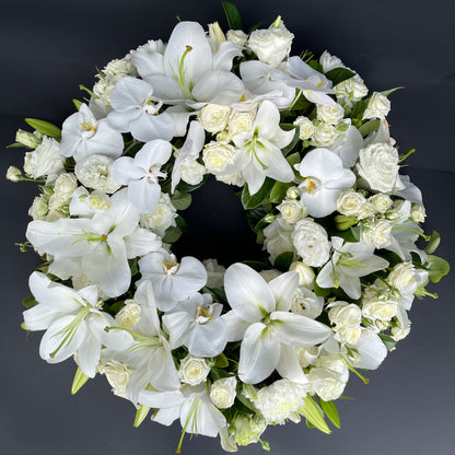 The Remembrance Wreath from FlowersonNortonSt features a circular floral arrangement of white lilies, roses, and orchids, creating a stunning tribute with green leaves scattered against a dark backdrop.