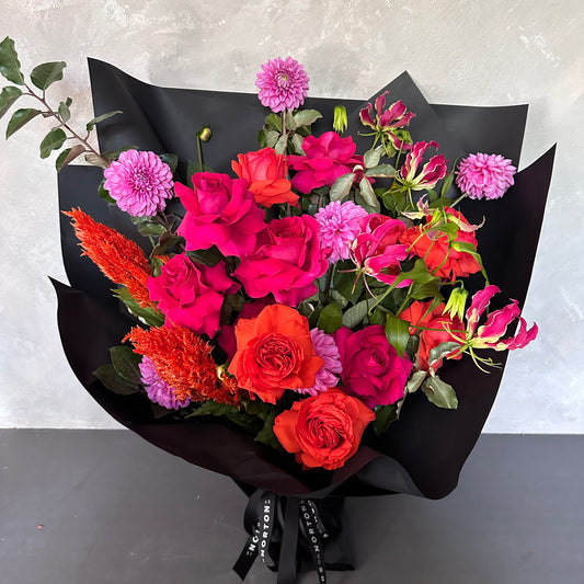 The Florist Choice - Red Nina Bouquet by FlowersonNortonSt features vibrant Red Nina and Colombian roses, bright pink blooms, and lush green foliage, elegantly wrapped in black paper with a black ribbon, set on a light gray backdrop.