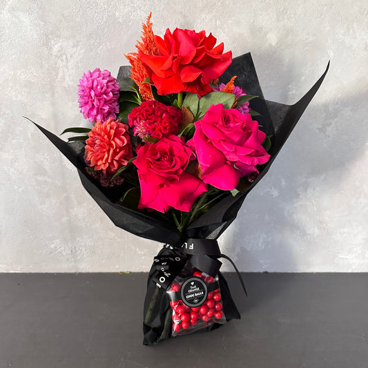 The Strawberry Kisses Mixed Bouquet 2024 by Flowers on Norton St, wrapped in black paper, features red and pink roses, dahlias, and a vibrant orange bloom. It includes a small package of Strawberry Kisses with a black ribbon against a light gray textured wall.