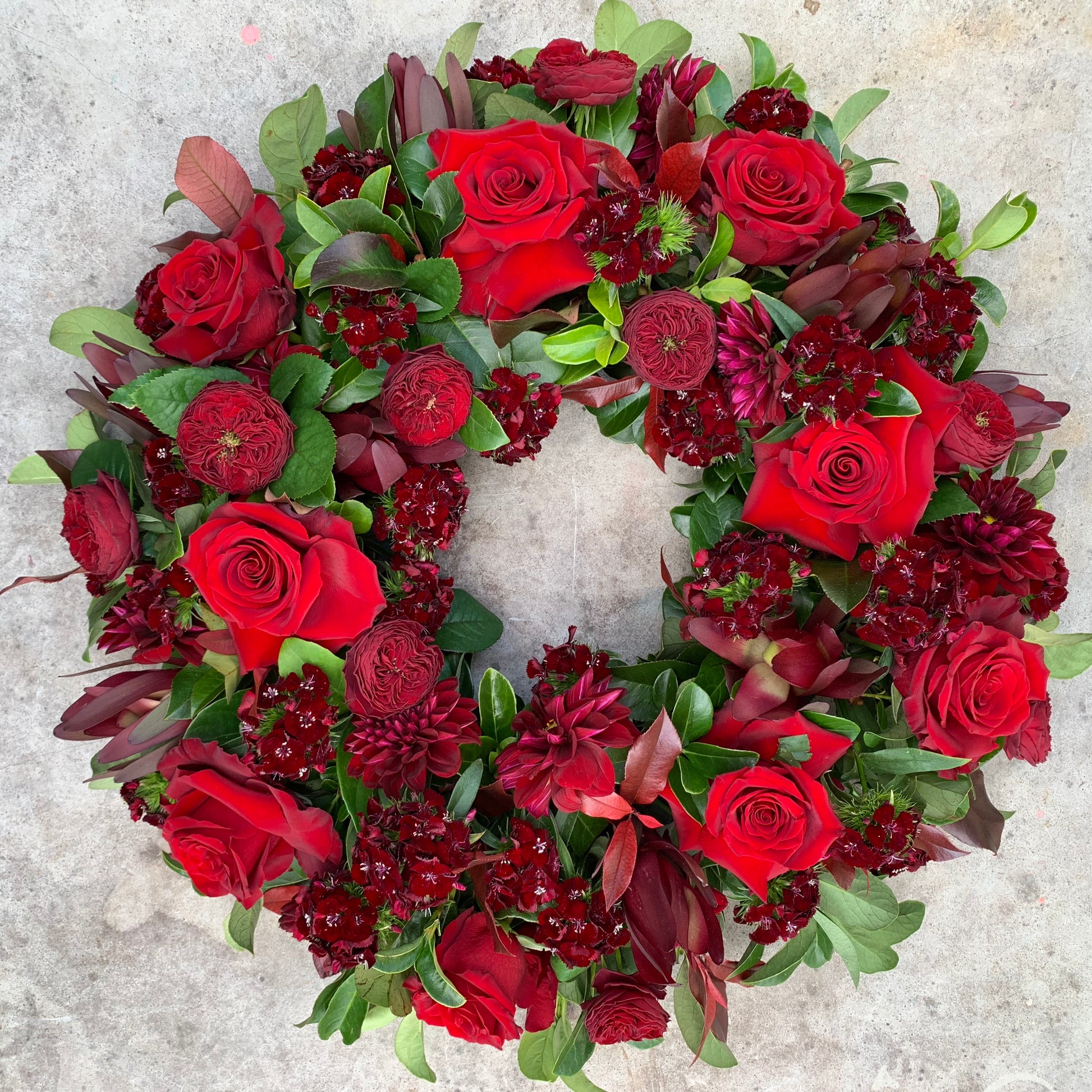 The Remembrance Wreath by FlowersonNortonSt beautifully displays vivid red roses, deep burgundy blooms, and lush green foliage on a light gray background, creating an ideal tribute.