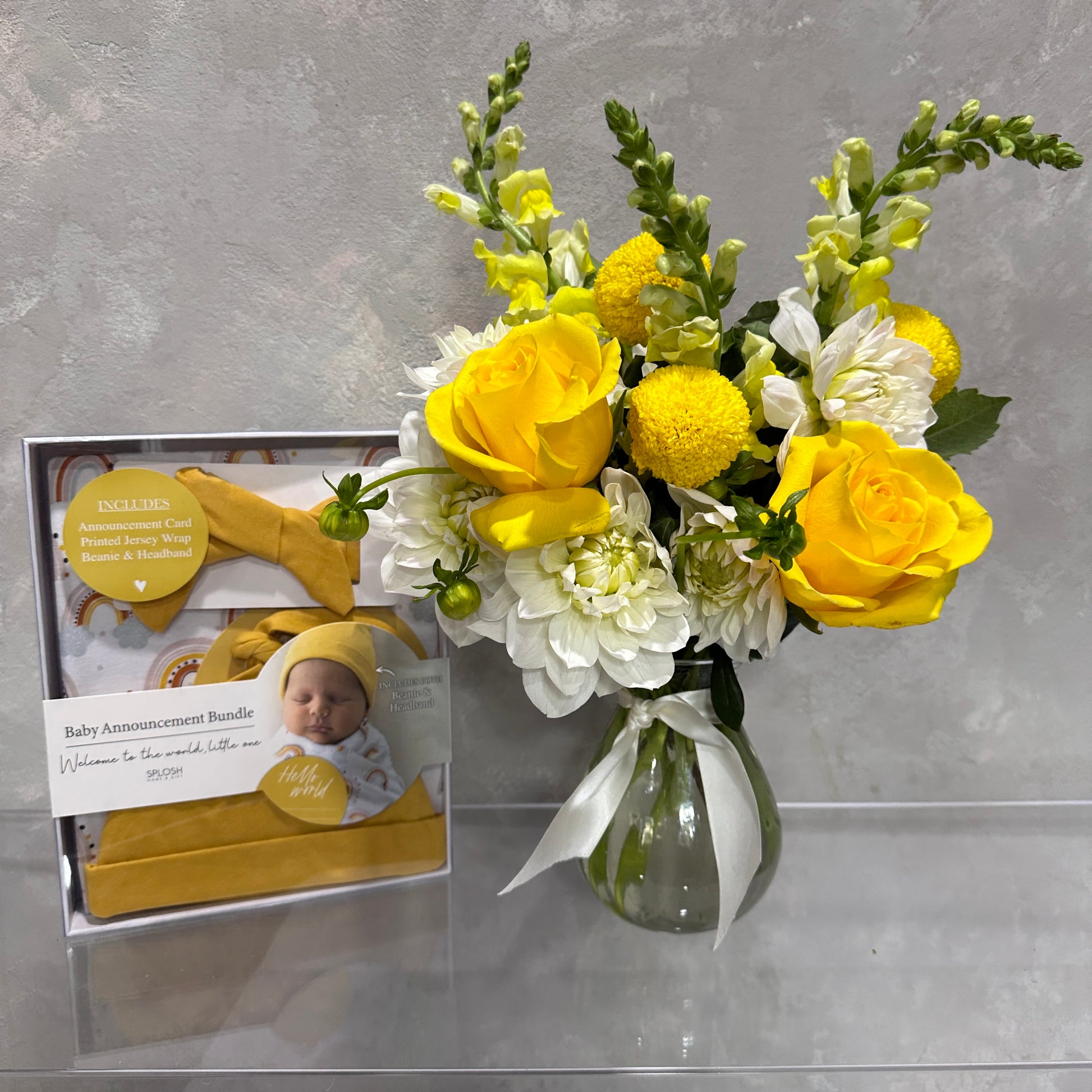 A vintage vase contains a bouquet of yellow roses and white flowers on a shelf. Beside it sits a "Welcome Baby" gift set from FlowersonNortonSt, featuring a photo of the newborn and yellow accessories, all beautifully arranged against a light gray wall.