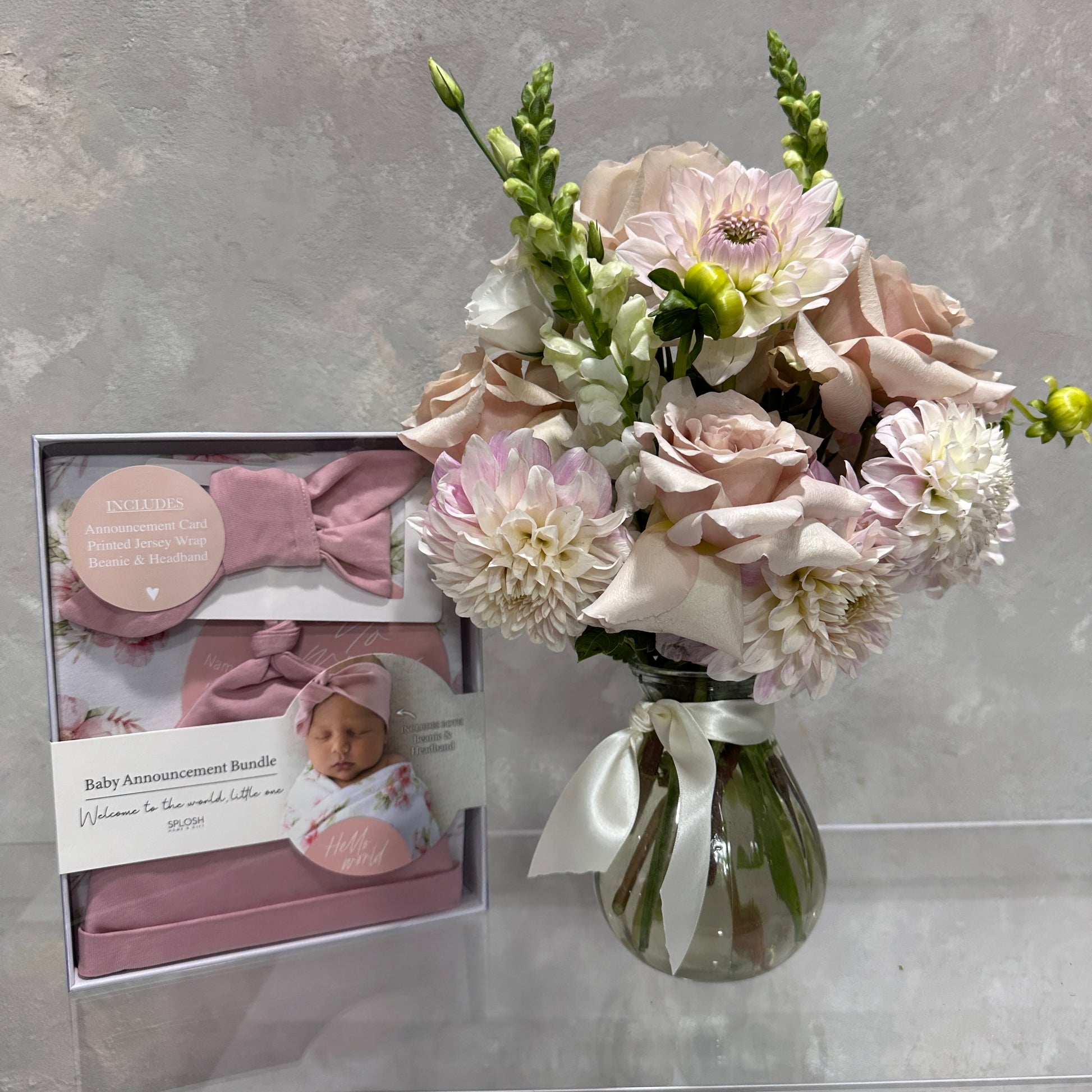 A delightful pink and white floral arrangement in a vintage vase sits beside the charming "Welcome Baby" gift set by FlowersonNortonSt. The set includes a pink outfit, matching headband, and a box featuring a baby photo with the message, "Welcome Baby! Welcome to the world, little one.