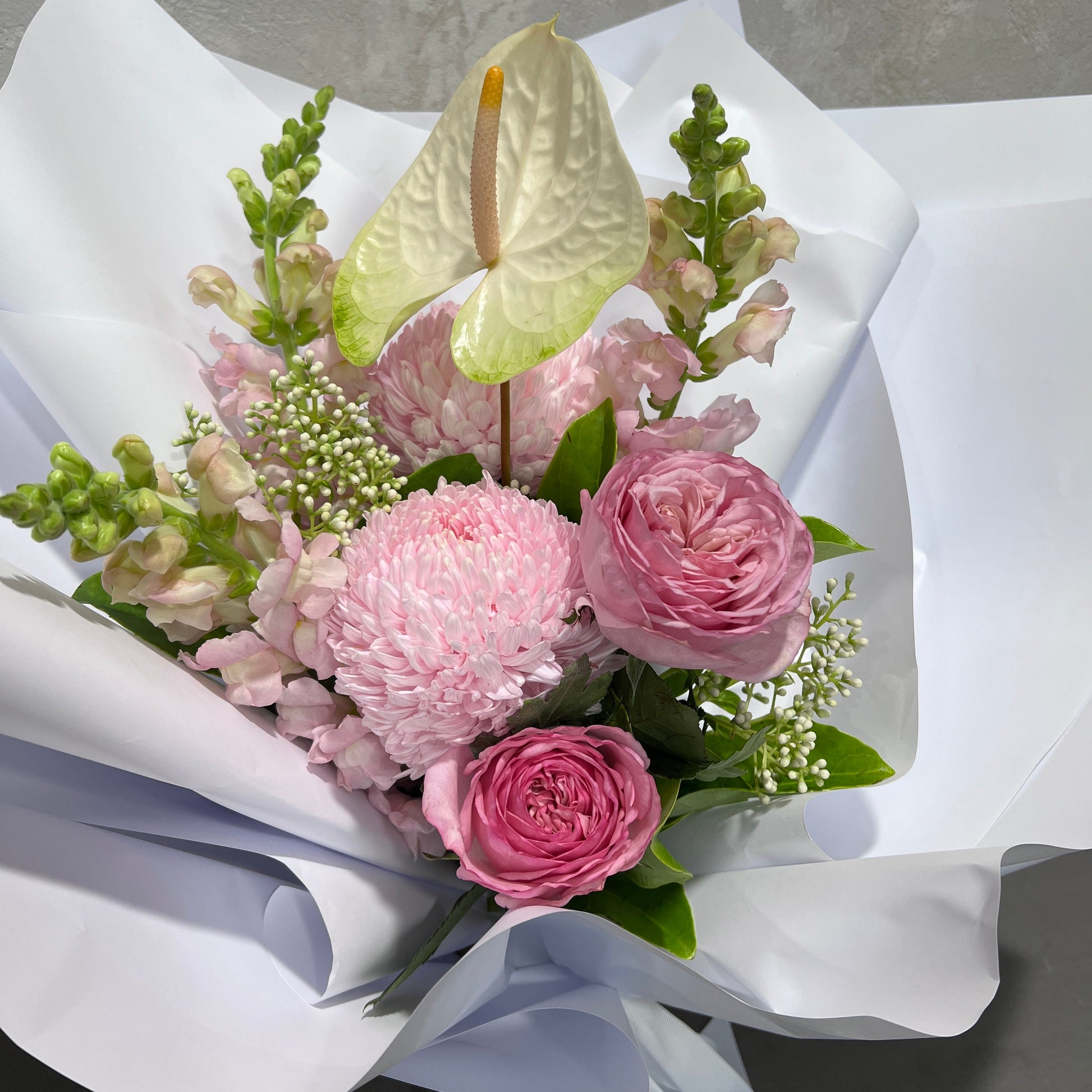 Presenting the "Tickle me Pink" by FlowersonNortonSt, a handcrafted botanical bouquet that showcases pink roses, a white anthurium, pink chrysanthemums, and small white blossoms. Elegantly wrapped in refined white paper, this distinctive gift perfectly embodies your unique style.