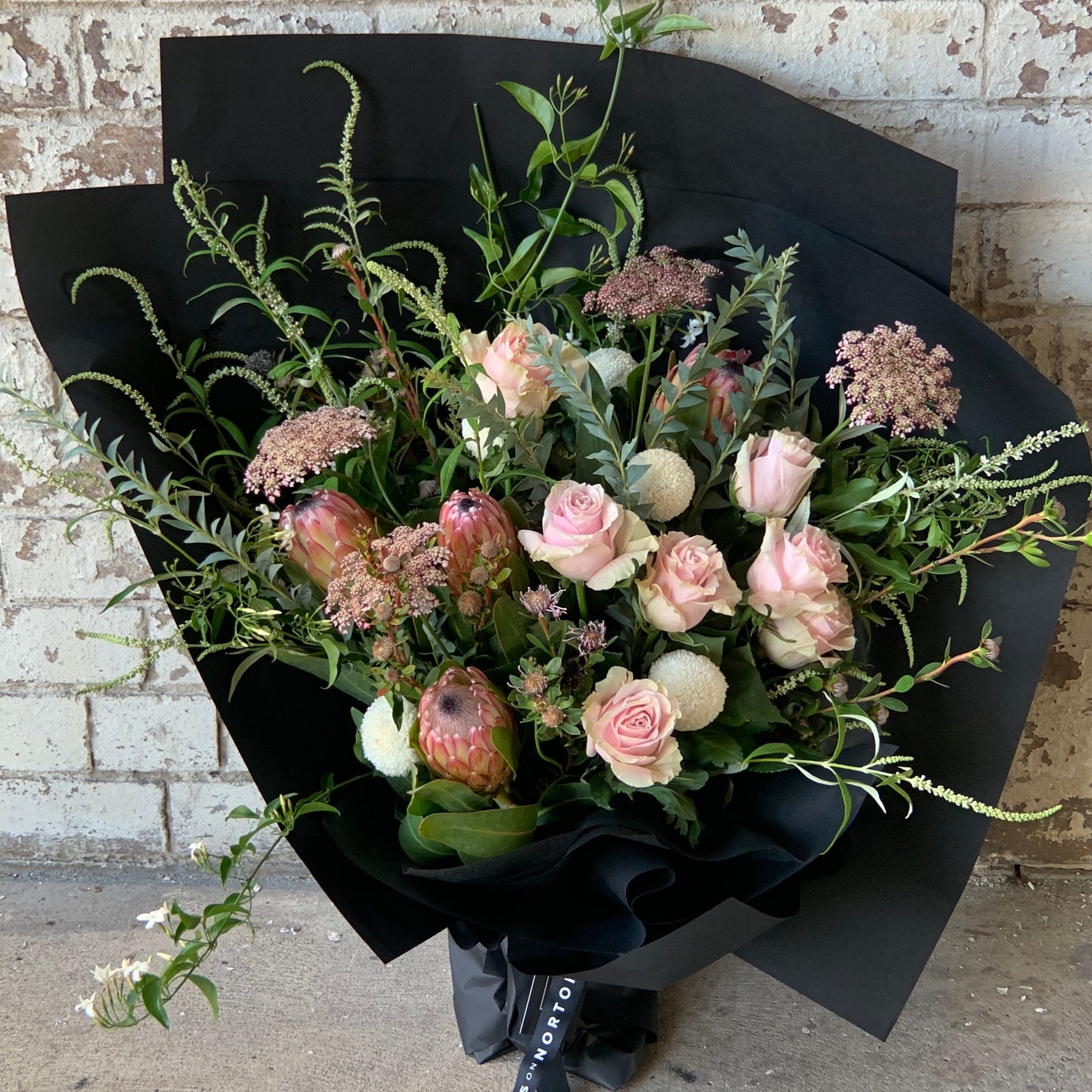 The "Delicate Roses + Natives" bouquet by Flowers on Norton St features a harmonious blend of delicate roses, native flowers, and proteas complemented by lush greenery, all elegantly wrapped in black paper against a rustic, weathered brick wall.