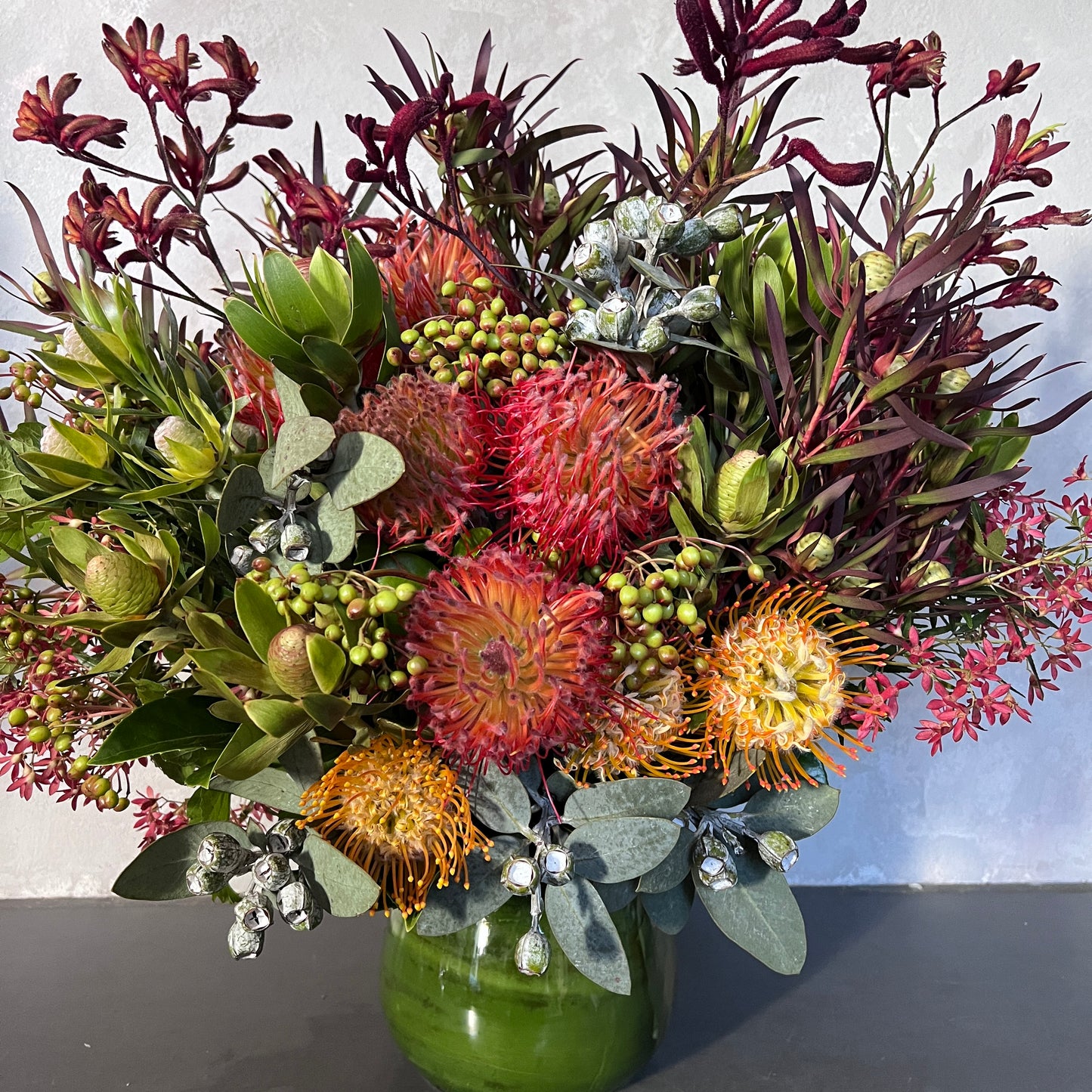 The "Wangal" by FlowersonNortonSt is a vibrant floral arrangement in a sculptural vase, featuring Australian natives like red kangaroo paws and orange pincushion proteas, complemented by lush tropical foliage and small clusters of berries against a light background.