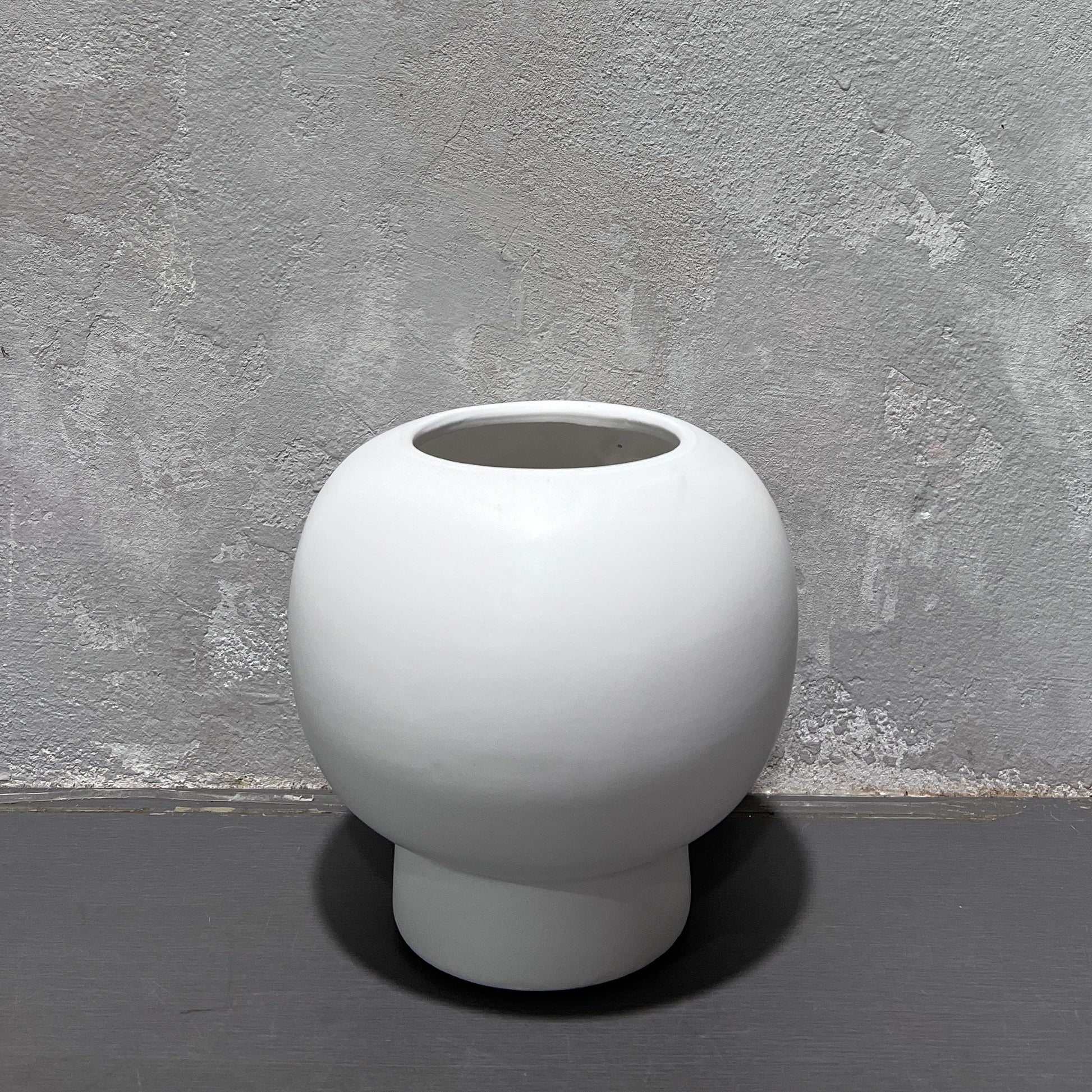 The Flowers on Norton St Bubble Vase, with its simple white ceramic design, rounded shape, and small opening, sits on a textured gray surface against a rough, light gray wall exhibiting subtle shade variations.