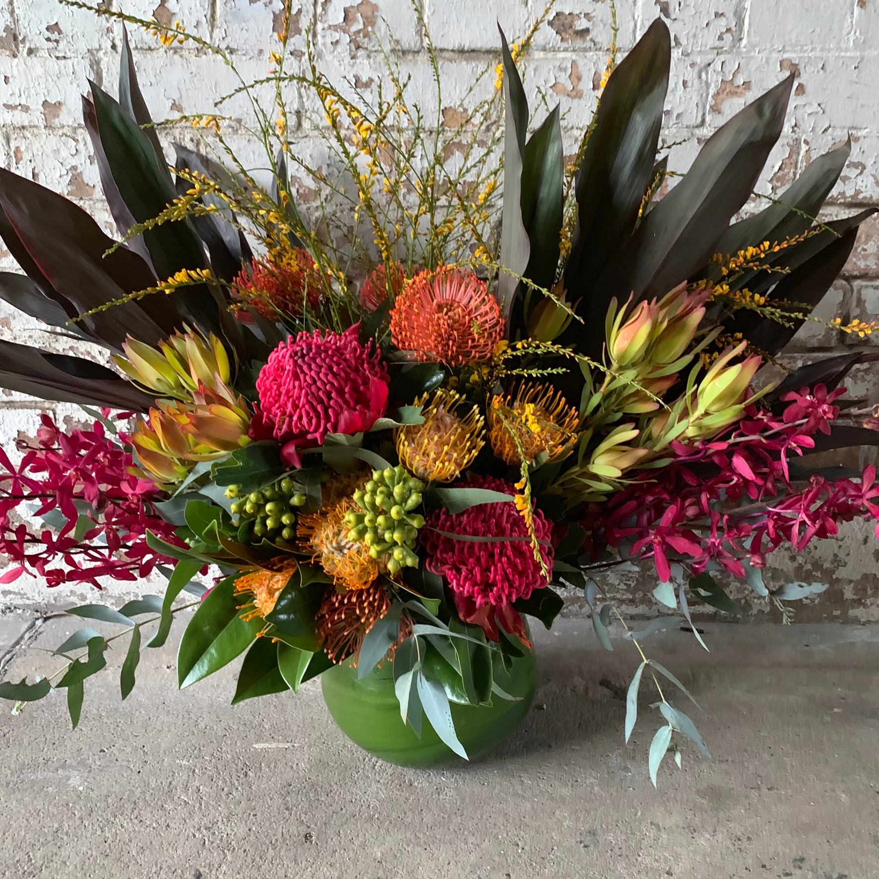 Introducing the "Wangal" by FlowersonNortonSt: This vibrant floral arrangement showcases Australian natives, including orange proteas and pink cockscomb, beautifully presented in a sculptural green vase with red orchids and dark green tropical foliage. Perfectly set against a textured brick wall to enhance its stunning visual appeal.