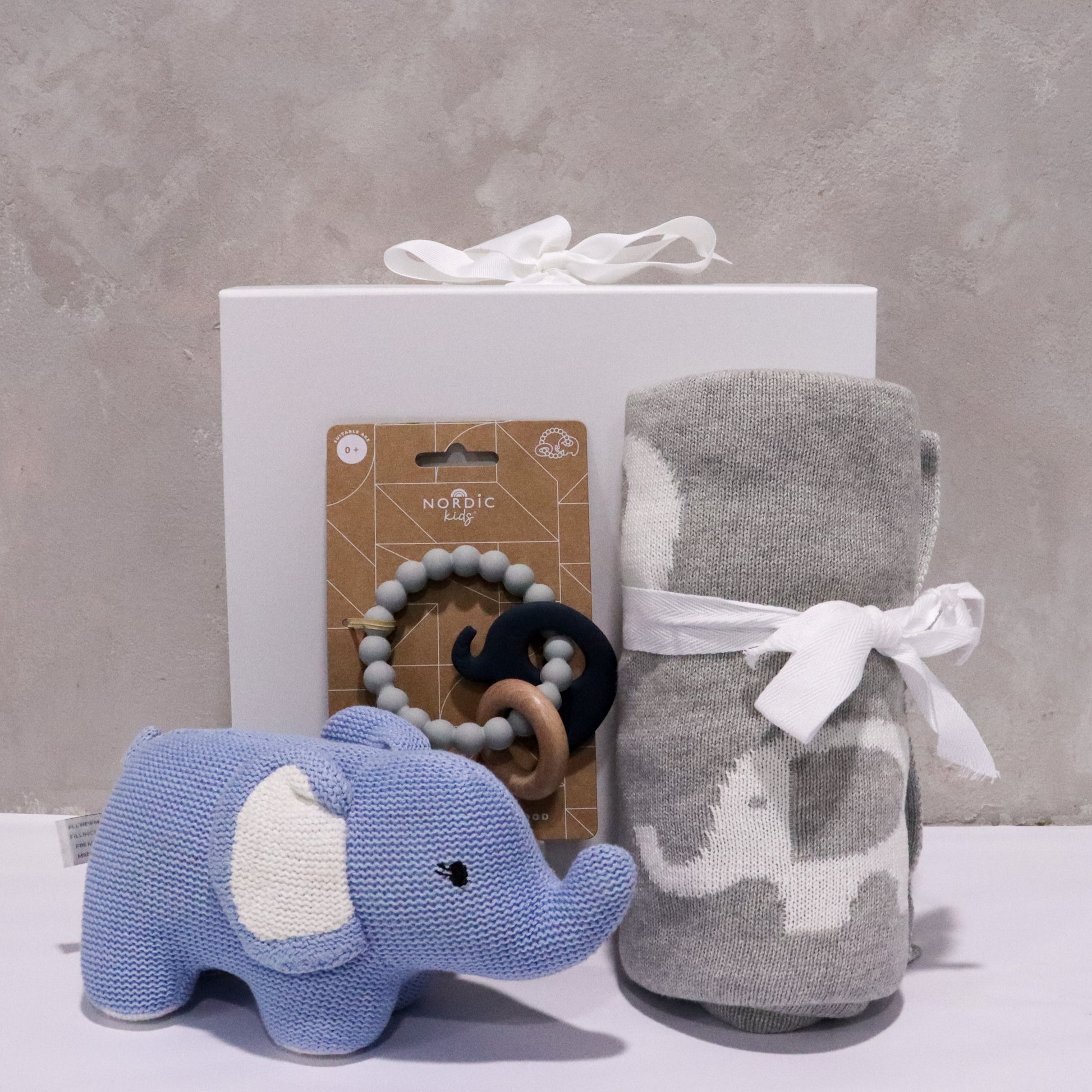 The Baby Elephant Gift Box by FlowersonNortonSt includes a delightful gray box with a white bow, a cozy blue Baby Elephant Plush, and a stylish baby blanket tied with a ribbon. The collection is completed by the Nordic Baby Teether made of wooden and silicone beads, all beautifully displayed against a concrete wall backdrop.