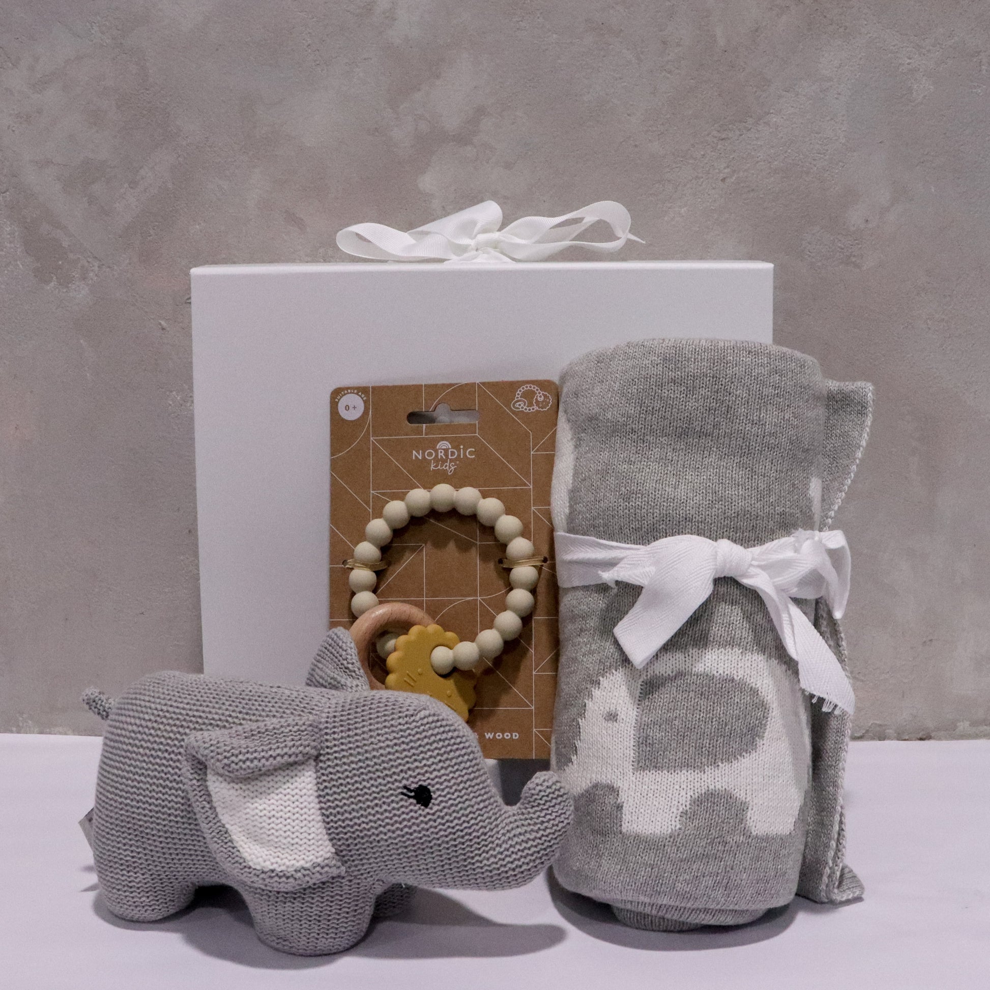 A Baby Elephant plush toy, along with a gray and white baby blanket featuring an elephant pattern, and a beige Nordic Baby Teether with decorative beads are tastefully arranged in front of a white gift box. The backdrop is an elegantly textured gray wall, adding to the calm ambiance of the presentation. This display showcases items from the FlowersonNortonSt Baby Elephant Gift Box collection.