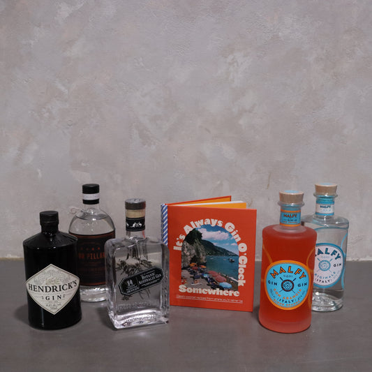 A collection of six gin bottles from Flowers on Norton St adorns the counter. At the center, a colorful booklet announces, "It's Always Gin O'Clock Somewhere." This set makes an ideal gift for gin enthusiasts, inviting them to indulge in timeless enjoyment.