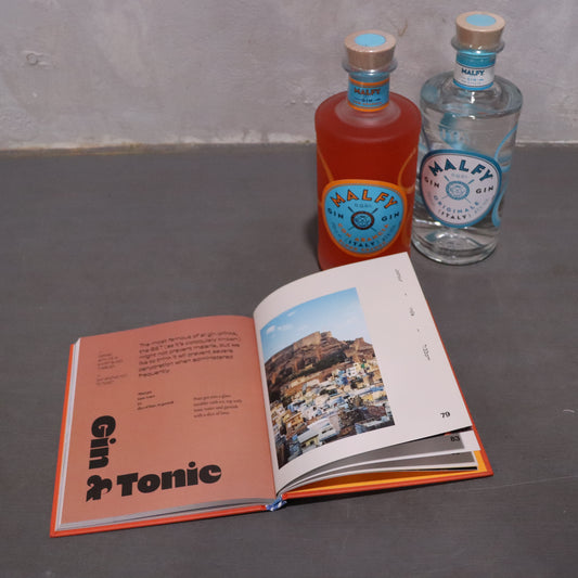 On a neutral gray table, an open cocktail recipe book sits beside two Flowers on Norton St gin bottles in pink and blue. The page titled "Gin & Tonic" displays a scenic view. This Gin Gift Set is the perfect present for enthusiasts seeking vibrant flavors, showcasing the allure of Blood Orange Gin.