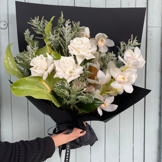 The Winter White Botanical Seasonal Bouquet by FlowersonNortonSt features white roses, cymbidium orchids, and green foliage elegantly wrapped in black paper. A hand displays the lush white and green arrangement against a light blue wooden backdrop.