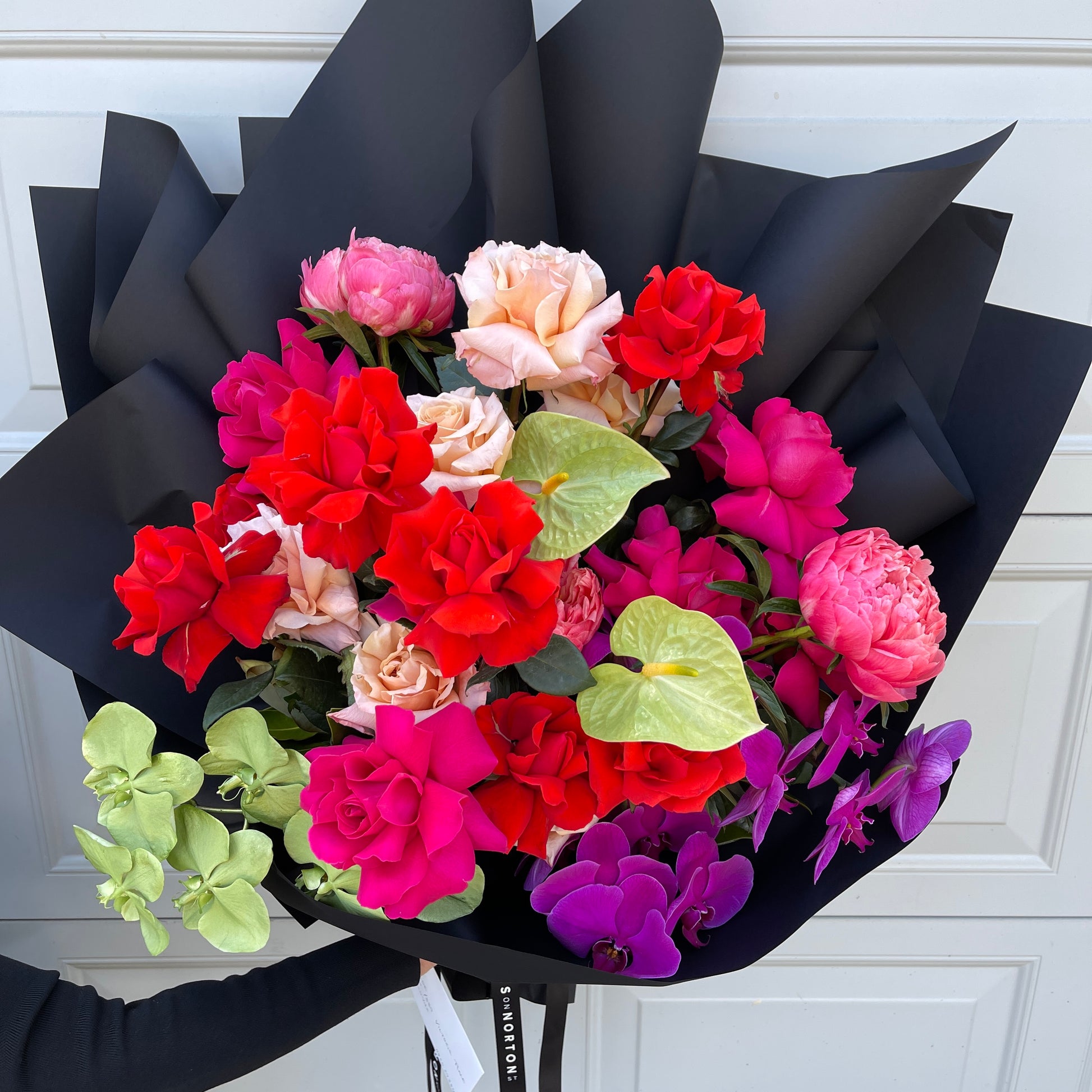 Introducing "Hello Lovely" from FlowersonNortonSt: a vibrant arrangement showcasing an exquisite blend of red, pink, and peach roses intricately combined with exotic tropical anthuriums and striking purple orchids, all gracefully wrapped in black paper set against a light grey backdrop.