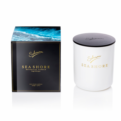 A white candle with a black lid and gold text, labeled "Sohum Gardenalia Candles," crafted from Eco Wax, stands next to its black box packaging featuring an ocean wave design and the brand name, "Flowers on Norton St," in gold. It brings a touch of serene floral fragrances to your space.