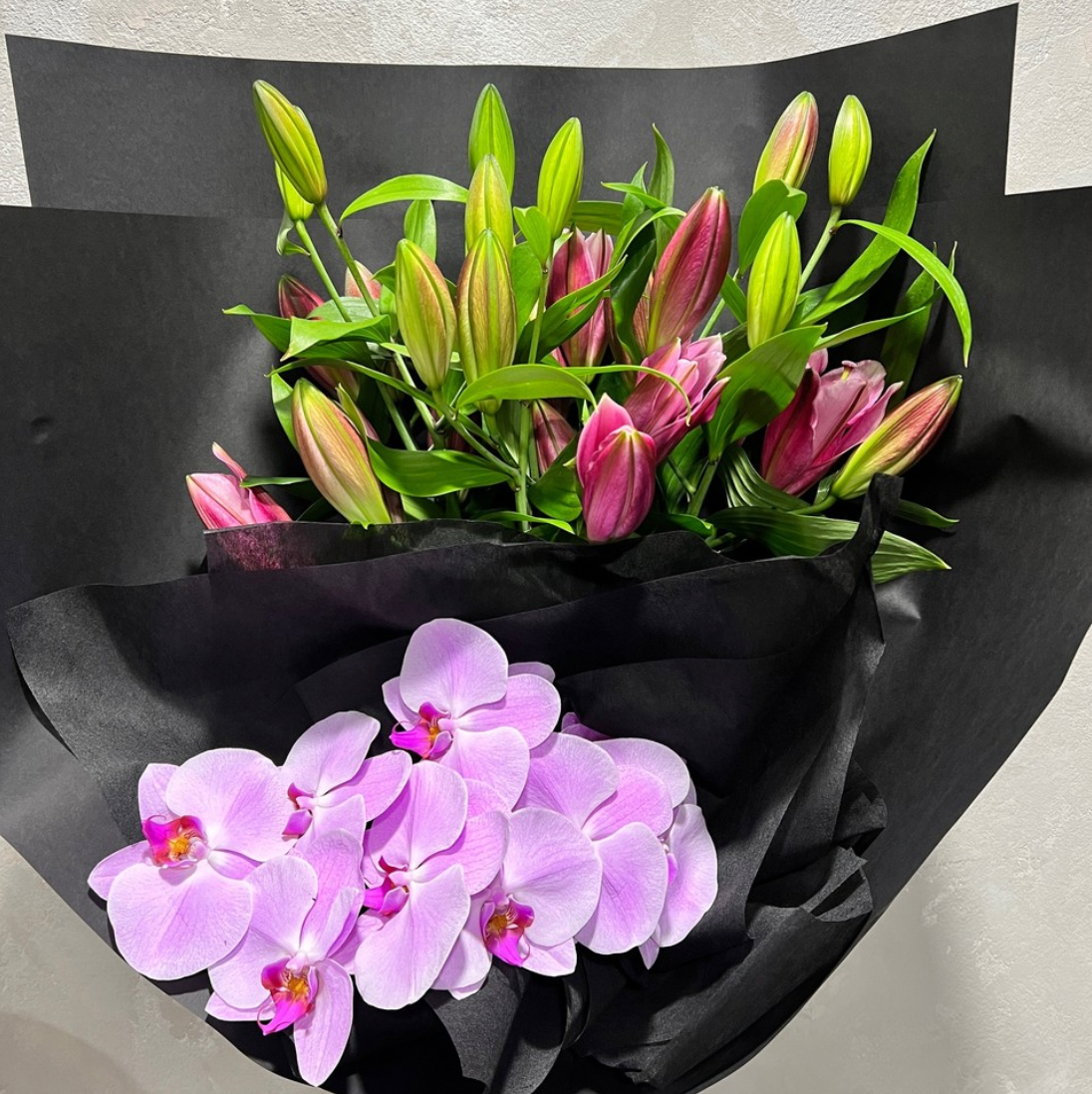 The "Colour Me - Pink" bouquet by Flowers on Norton St features seasonal pink flowers, including unopened lilies and light purple orchids, beautifully arranged against a black background—making it the perfect romantic gift.