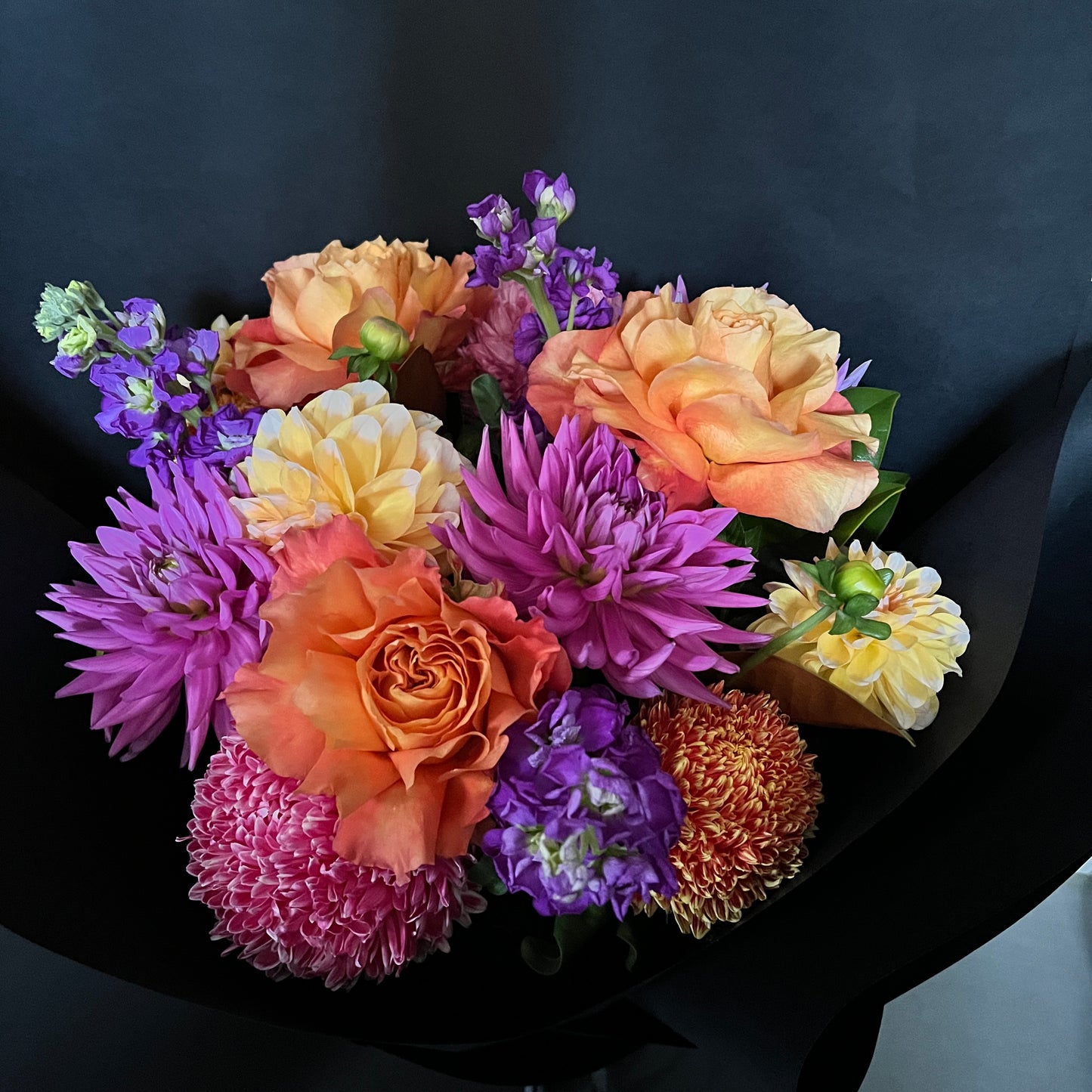 A vibrant bouquet by FlowersonNortonSt against a black background highlights the contemporary use of color with seasonal flowers, showcasing orange roses, purple chrysanthemums, yellow dahlias, and small purple lilacs. The striking contrast creates a captivating winter warmer posy.