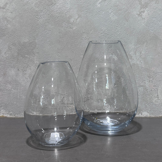 A pair of Teardrop Glass Vases from Flowers on Norton St are arranged together on a gray surface, set against a textured gray wall. Their smooth, curved surfaces catch the light beautifully.