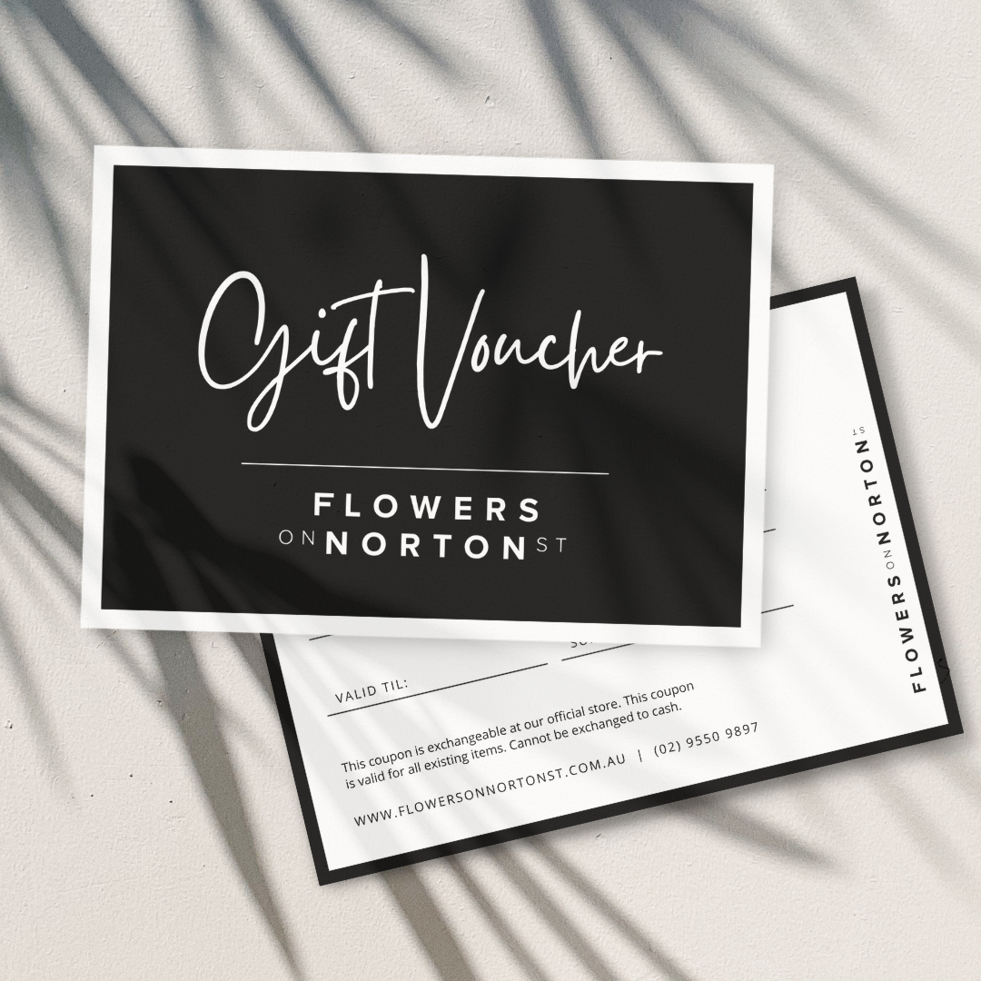 A chic Flowers on Norton St Gift Voucher rests elegantly on a light surface, adorned with leafy shadows. This black and white voucher outlines details about its validity and exchange conditions, making it the perfect token for flowers or exquisite homewares.