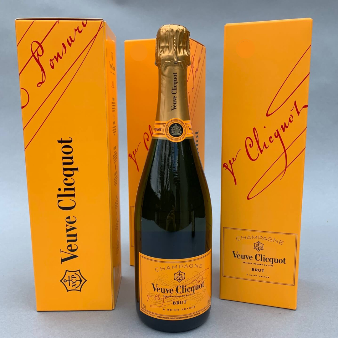 A bottle of Veuve Clicquot from FlowersonNortonSt stands proudly in front of two vibrant orange boxes. The labels showcase elegant script and branding, set against a neutral gray backdrop.