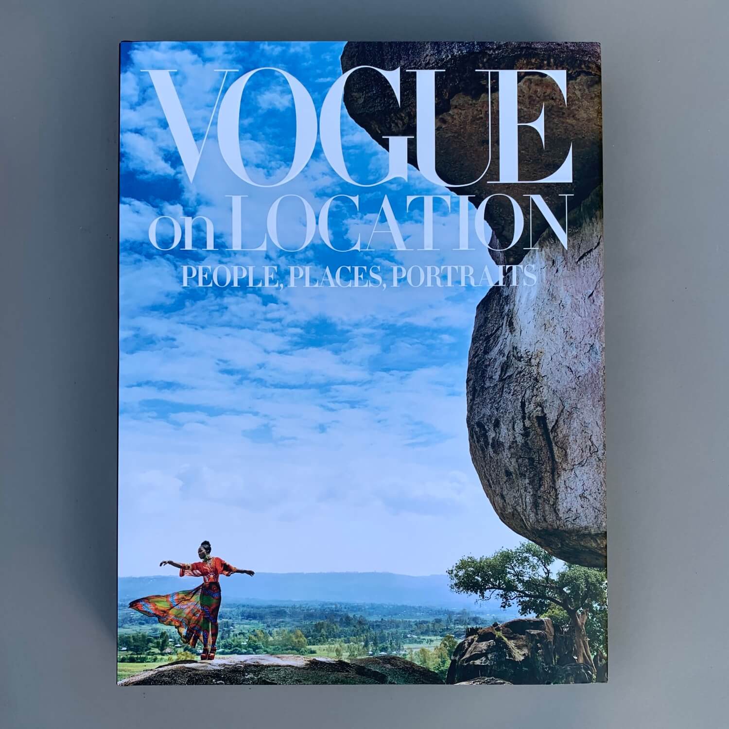 The book cover for "Vogue on Location" by FlowersonNortonSt features travel fashion, depicting a person in vibrant clothing standing atop a rock formation against an expansive landscape and sky.