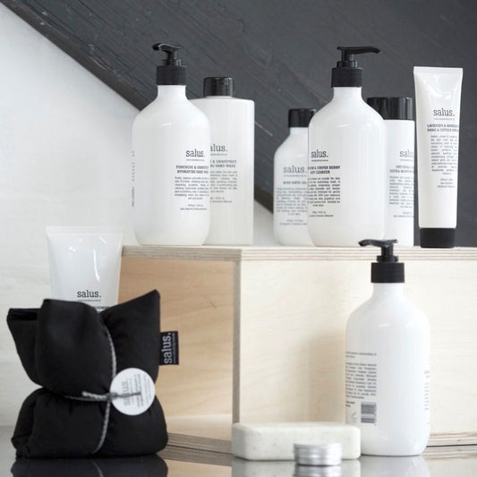 A selection of FlowersonNortonSt skincare products, featuring the Salus Hand Wash, is elegantly showcased on wooden blocks. The white bottles and tubes are adorned with black text and caps. Completing the minimalist arrangement are a black fabric pouch and a silver tin, with essential oils subtly enhancing the ambiance.