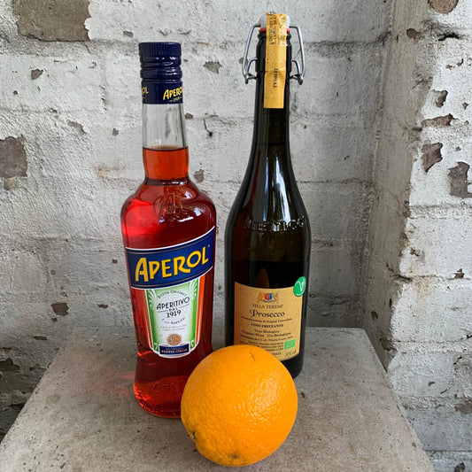 An Aperol Spritz Gift Pack by FlowersonNortonSt, featuring a bottle of Aperol, a bottle of Prosecco, and an orange arranged on a stone surface against a brick wall, captures the vibrant charm of an Aperol Spritz.