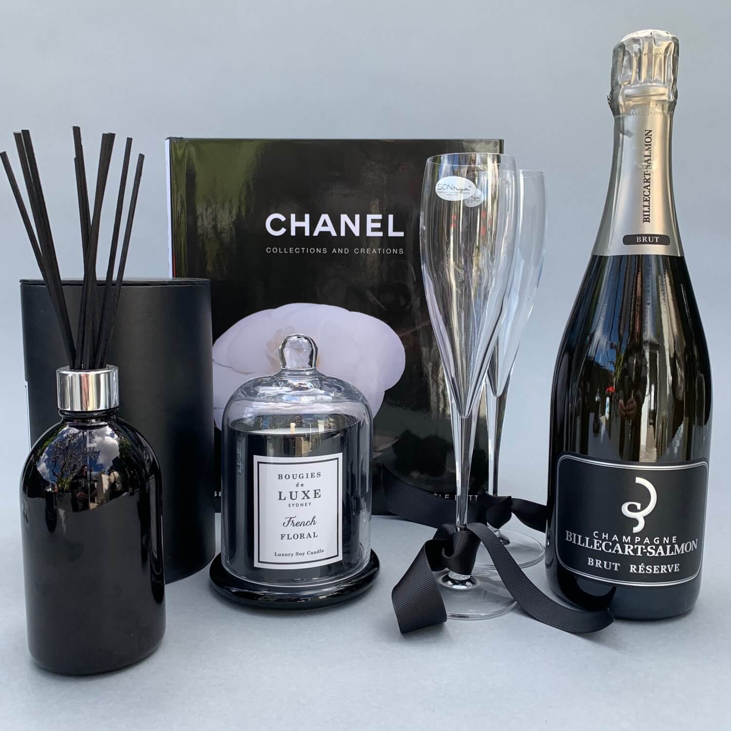 The Chanel Luxury Gift Box by FlowersonNortonSt features a Chanel Collections and Creations book, a black diffuser, a candle in a glass holder, two elegant champagne flutes, and Billecart-Salmon Brut Reserve champagne—all elegantly set against a soft gray background.