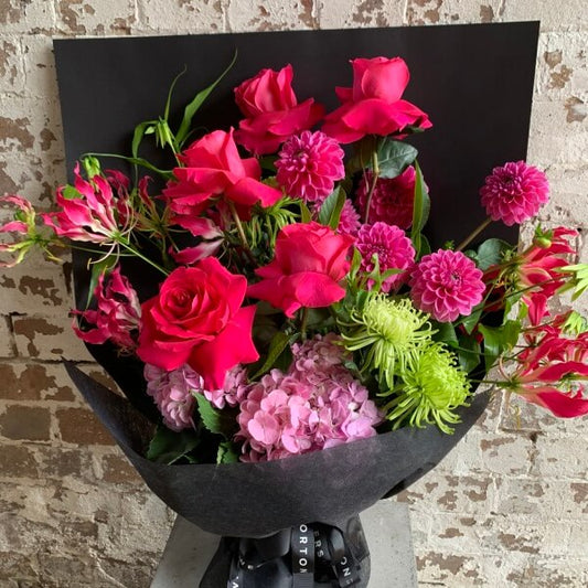 The Summer Splice Pink Bouquet by FlowersonNortonSt showcases a stunning collection of vibrant pink roses, Colombian roses, dahlias, and pink hydrangeas. Enhanced with green chrysanthemums and exotic flame lilies, this bouquet is elegantly presented in dark wrapping paper and makes a striking statement against a rustic brick wall background.